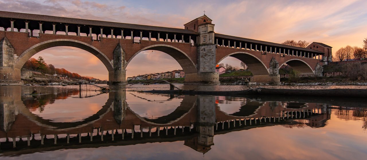Pavia’da gün batımında Ticino Nehri üzerindeki üstü kapalı Ponte Coperto ve sudaki yansıması